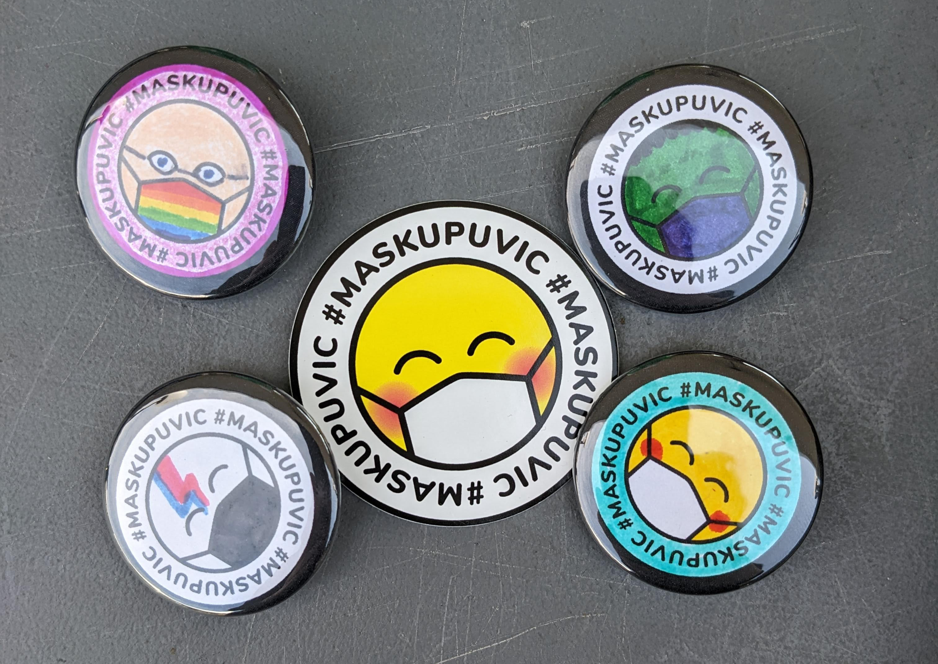 A #MaskUpUVic sticker in yellow surrounded by 4 custom coloured pins: the hulk, yellow and teal, David Bowie, and pride mask with glasses.